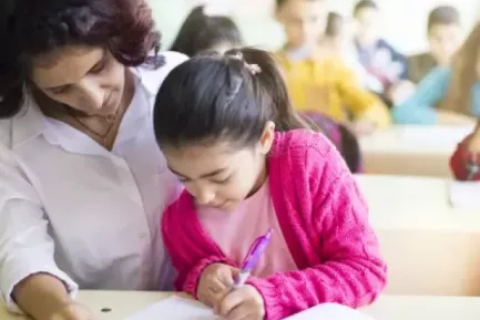A teacher sits close to a child, who is deeply concentrating on writing in a notebook.